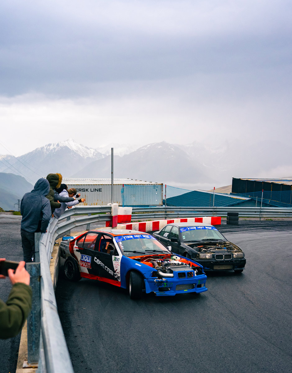 Image montrant deux voitures en train de faire un drift près du mur sur le circuit d'Andorre, avec les montagnes Pyrénées enneigées, lors d'une compétition de drift.